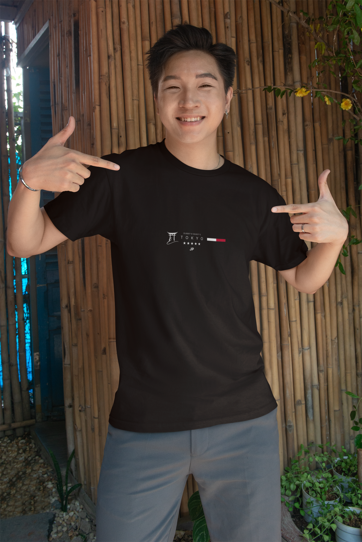 Smiling man standing in front of a bamboo wall, pointing with both hands at his black "TOKYO" t-shirt, which features a minimalist city-themed design in white and red; wearing grey pants and a silver bracelet, in a casual outdoor setting with potted plants.