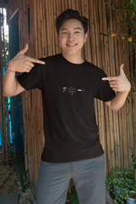Smiling man standing in front of a bamboo wall, pointing with both hands at his black "TOKYO" t-shirt, which features a minimalist city-themed design in white and red; wearing grey pants and a silver bracelet, in a casual outdoor setting with potted plants.