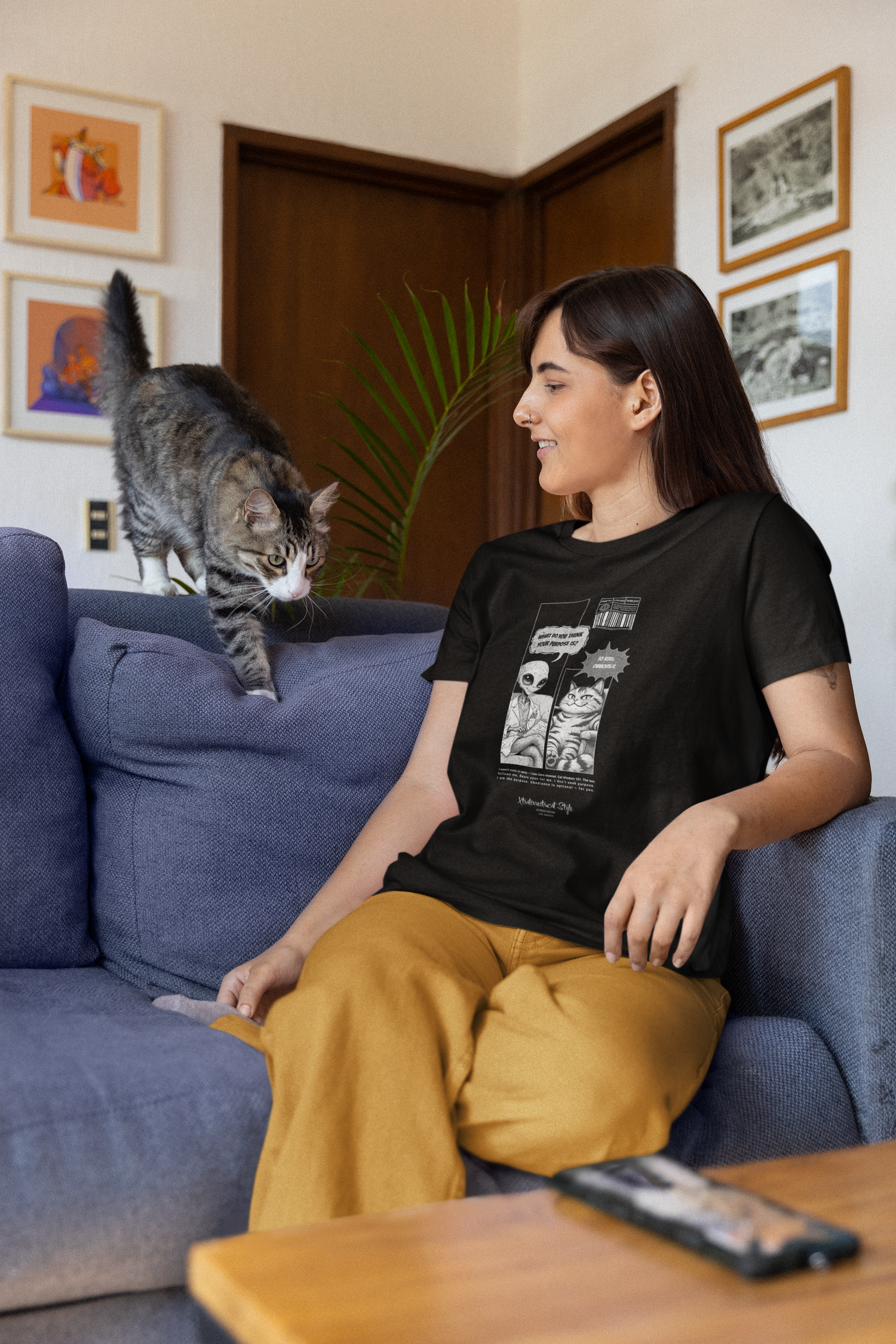 A woman sitting on a blue sofa, wearing a black "To Rule" graphic tee featuring a comic-style alien and cat dialogue. The alien asks, “What do you think your purpose is?” and the cat responds, “To rule. Obviously.” A grey tabby cat walks across the sofa towards her. The setting is a cozy living room with wooden accents, framed art, and houseplants in the background.
