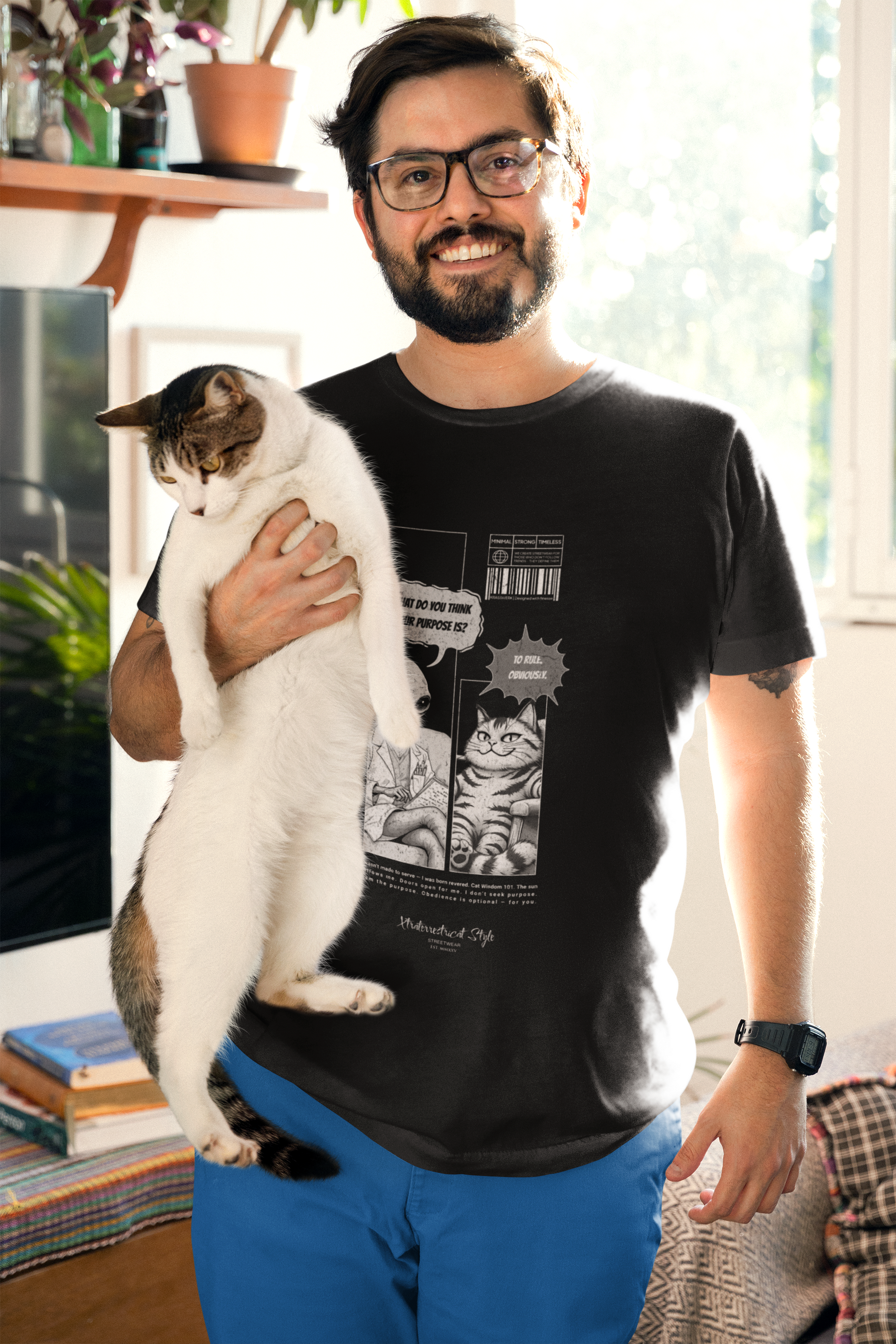A man with glasses and a beard smiles while holding a white-and-brown cat in his arm. He wears a black "To Rule" comic-style t-shirt featuring an alien asking “What do you think your purpose is?” and a cat confidently replying, “To rule. Obviously.” The scene is warmly lit by sunlight through a nearby window, with books and indoor plants in the cozy background.