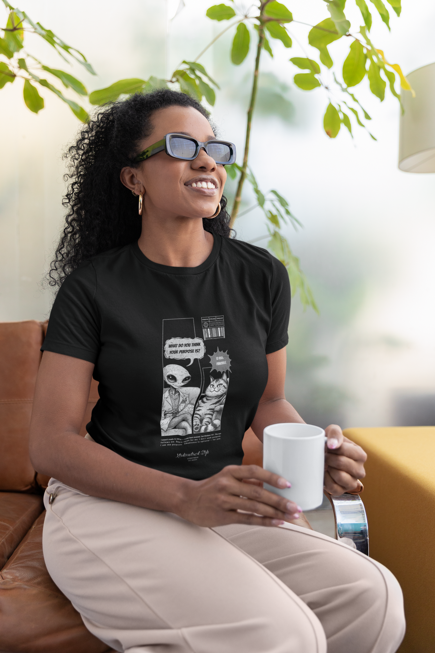 A woman with curly hair and sunglasses sits on a couch, smiling and holding a white mug. She wears a black "To Rule" graphic tee featuring a comic-style alien and a cat. The alien asks, “What do you think your purpose is?” and the cat replies, “To rule. Obviously.” A tall indoor plant and a yellow table complete the bright, modern background.