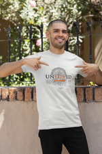 A smiling man points proudly at his white UNBOWED t-shirt, standing in front of a black iron fence with sunlit flowers in the background. The design features a tree with deep roots and the word “UNBOWED” in bold black and orange text.