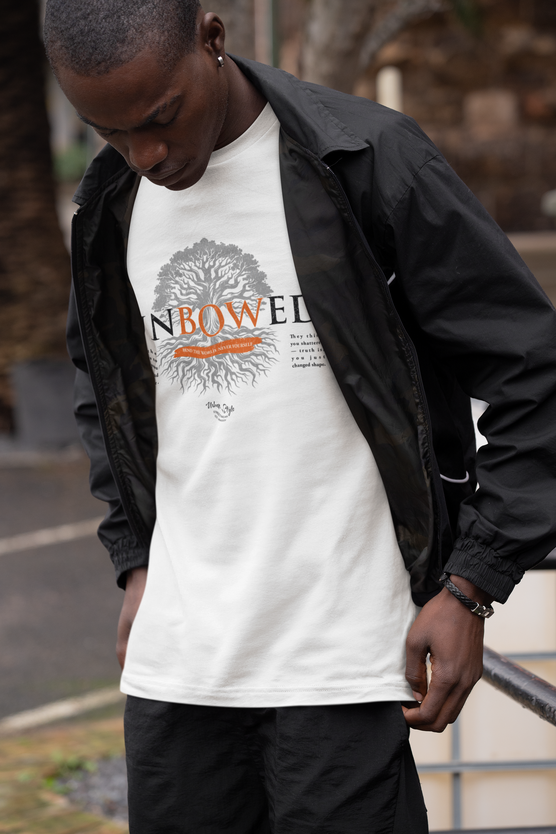 A man in a white UNBOWED t-shirt stands outdoors, looking down with quiet strength. His jacket is open, revealing the bold tree design and the word “UNBOWED” across his chest, accented by deep orange roots and lettering.