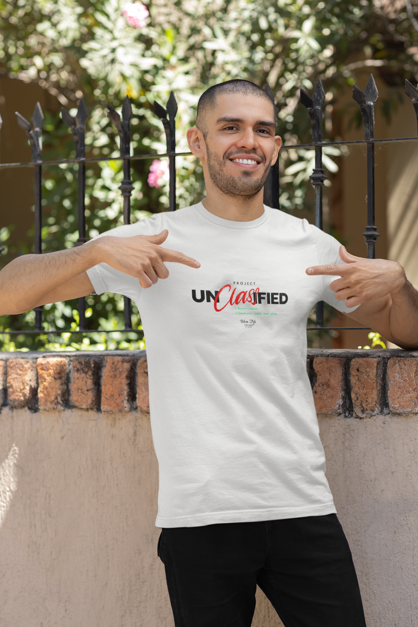 Smiling male model pointing at the white “PROJECT UNCLASSIFIED” t-shirt in a sunny courtyard. He leans casually against a brick wall, framed by black iron bars and vibrant greenery. With a confident grin, he highlights the bold black and red typography on the shirt — a quiet nod to secret missions and untold stories wrapped in streetwear attitude.