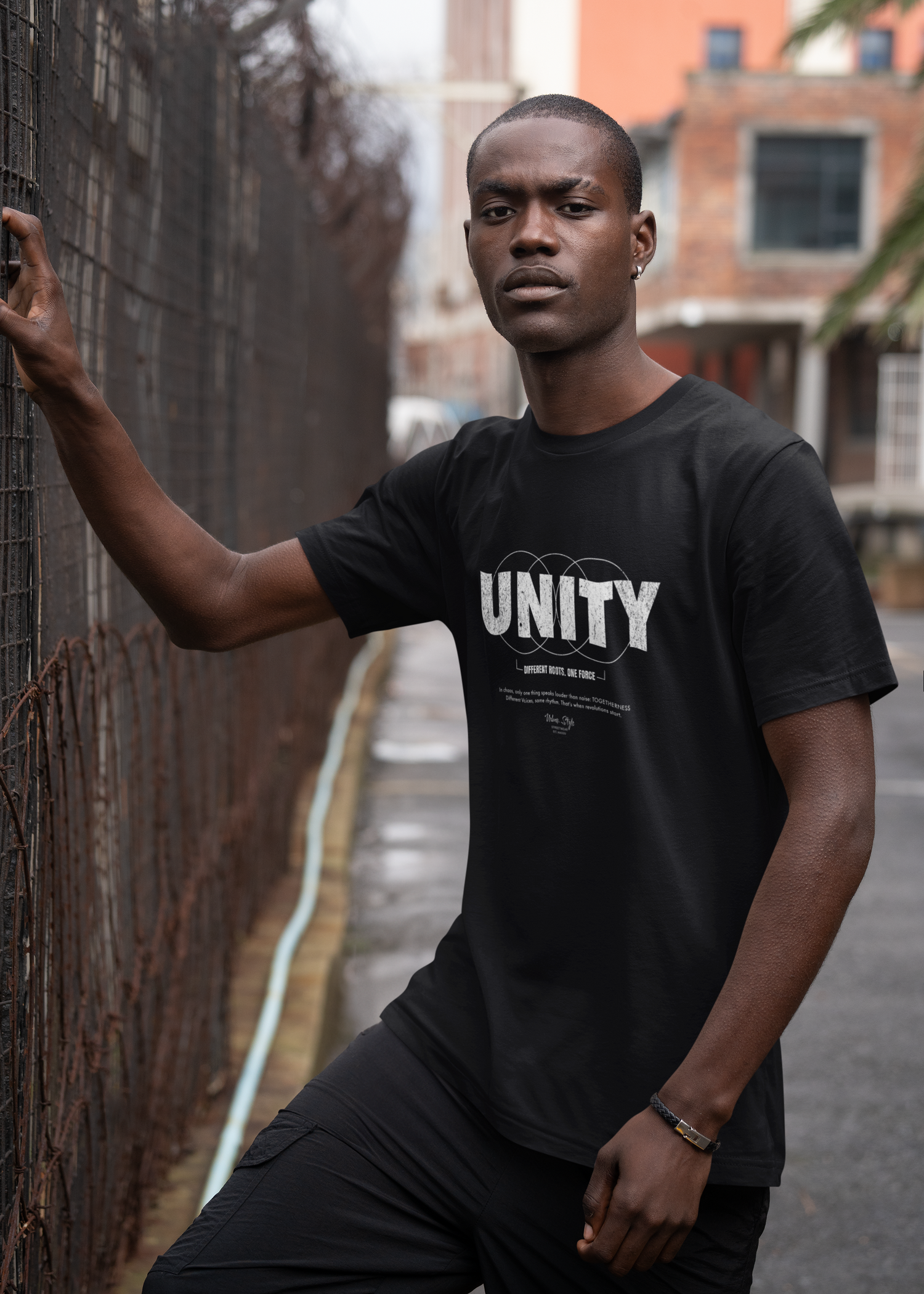 Model stands firmly beside a rusted fence, wearing the black UNITY t-shirt. His direct gaze and composed stance echo the shirt’s message — strength through solidarity, presence without noise. A portrait of power, pride, and unshakable unity.