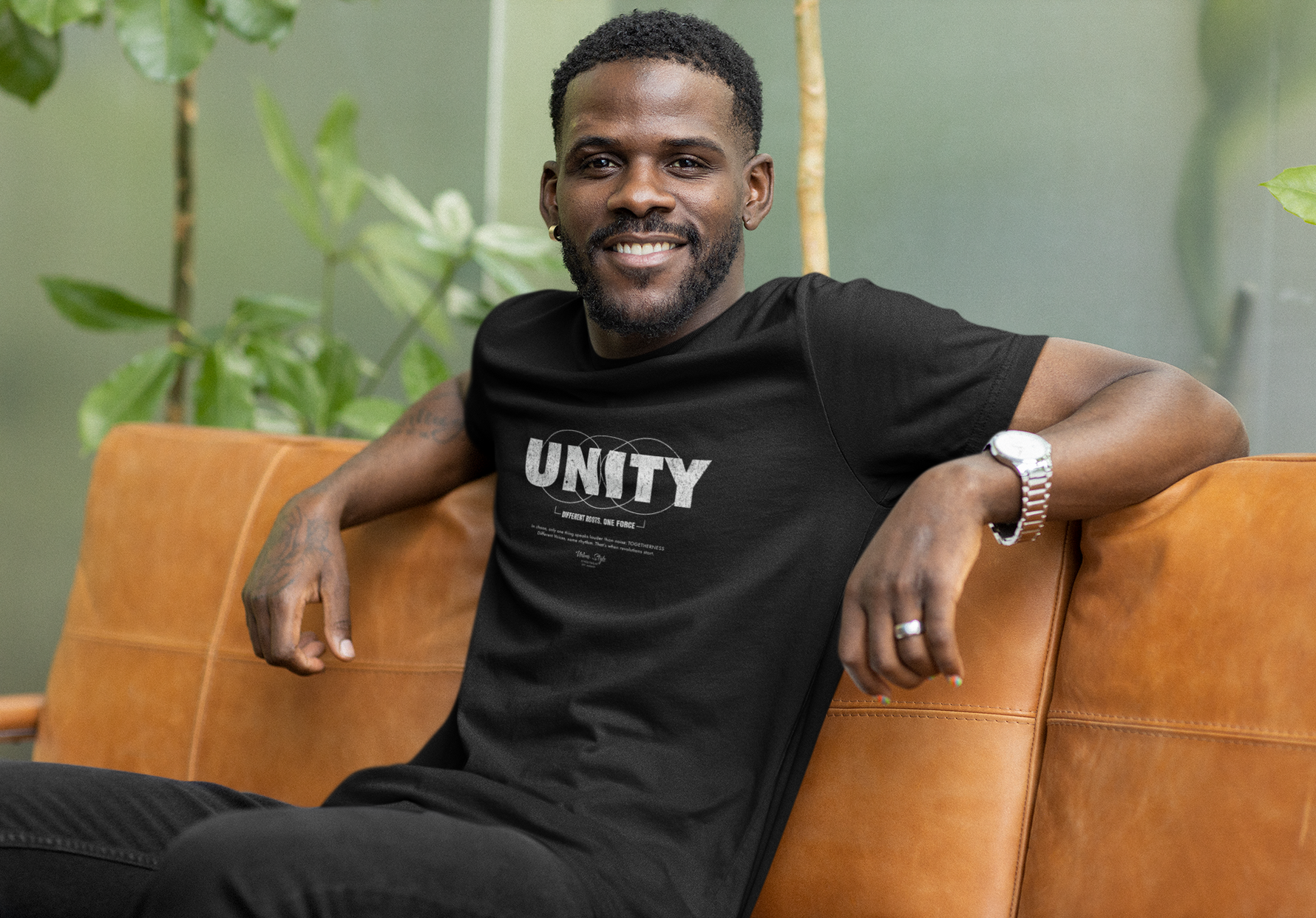 A confident man relaxes on a leather couch, smiling calmly in the UNITY t-shirt — a visual echo of strength through connection. Surrounded by warm tones and greenery, his posture radiates ease, but the message on his chest carries weight: different roots, one force.