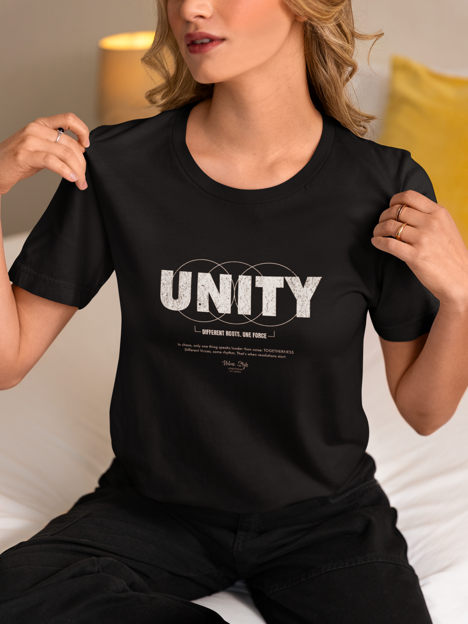 A woman seated on a cozy bed lifts her shirt slightly to reveal the UNITY design — a bold statement of harmony and purpose. Her relaxed confidence and subtle expression mirror the message: many voices, one vision. Togetherness isn't loud — it's powerful.