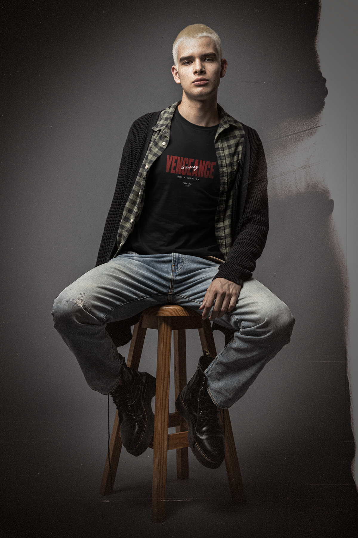 Young man with bleached blonde hair sits confidently on a wooden stool in a moody studio setting. He wears a black “VENGEANCE – Not a Solution” t-shirt layered under an open plaid shirt and oversized cardigan, styled with distressed jeans and combat boots. The gritty background and soft lighting enhance the rebellious, streetwear attitude of the KRASSWERK design. The bold red typography on the shirt stands out, emphasizing the message of silent strength and defiant energy.