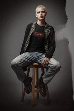 Young man with bleached blonde hair sits confidently on a wooden stool in a moody studio setting. He wears a black “VENGEANCE – Not a Solution” t-shirt layered under an open plaid shirt and oversized cardigan, styled with distressed jeans and combat boots. The gritty background and soft lighting enhance the rebellious, streetwear attitude of the KRASSWERK design. The bold red typography on the shirt stands out, emphasizing the message of silent strength and defiant energy.
