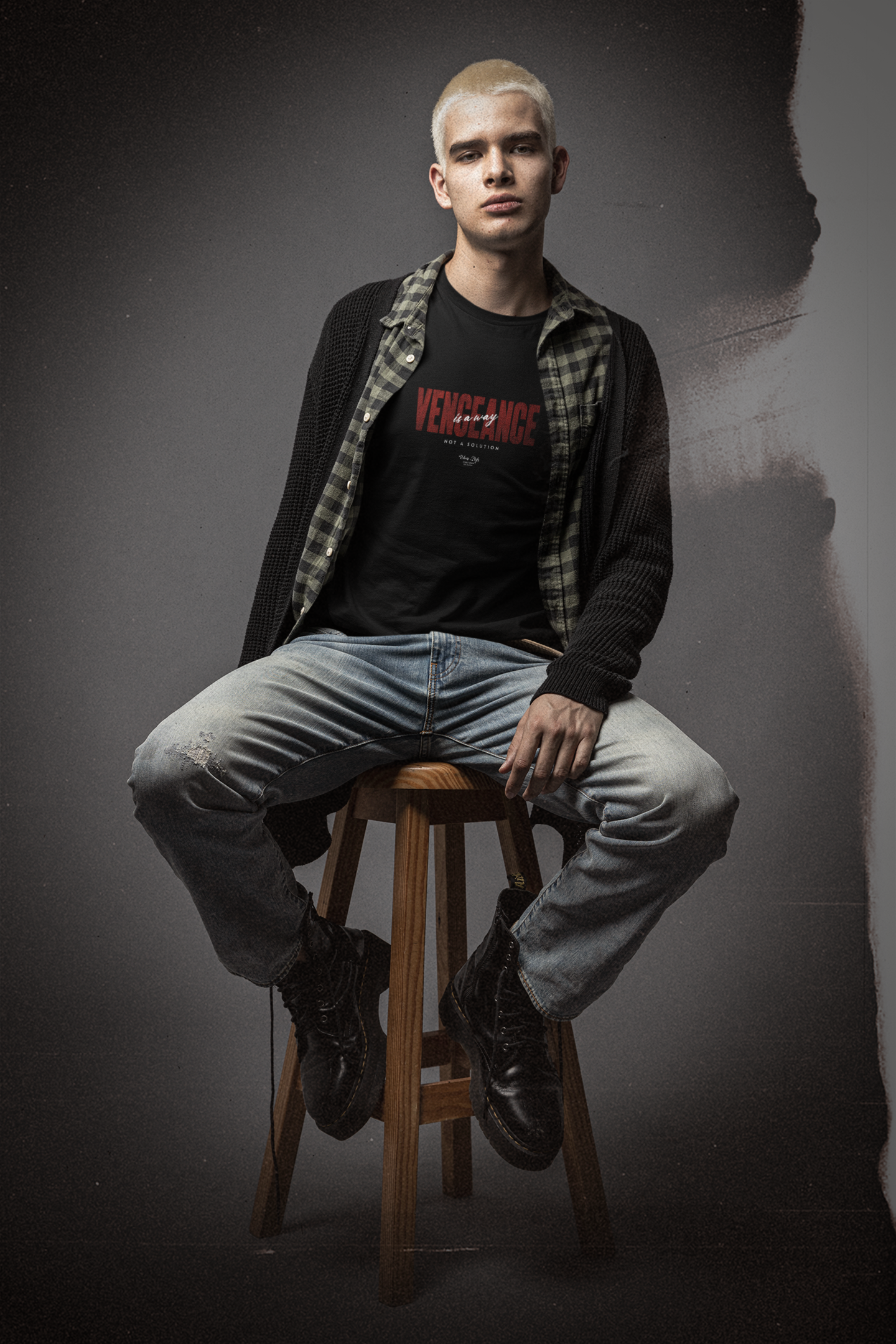 Young man with bleached blonde hair sits confidently on a wooden stool in a moody studio setting. He wears a black “VENGEANCE – Not a Solution” t-shirt layered under an open plaid shirt and oversized cardigan, styled with distressed jeans and combat boots. The gritty background and soft lighting enhance the rebellious, streetwear attitude of the KRASSWERK design. The bold red typography on the shirt stands out, emphasizing the message of silent strength and defiant energy.