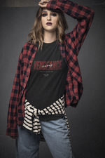Young woman with bold makeup poses with confidence, lifting one arm above her head in a strong stance. She wears a black “VENGEANCE — is a way, not a solution” t-shirt layered under an open red flannel shirt, with a striped sweatshirt tied around her waist. Her intense gaze and layered streetwear styling reflect a rebellious, unapologetic energy that embodies the KRASSWERK message of silent resistance and personal truth.