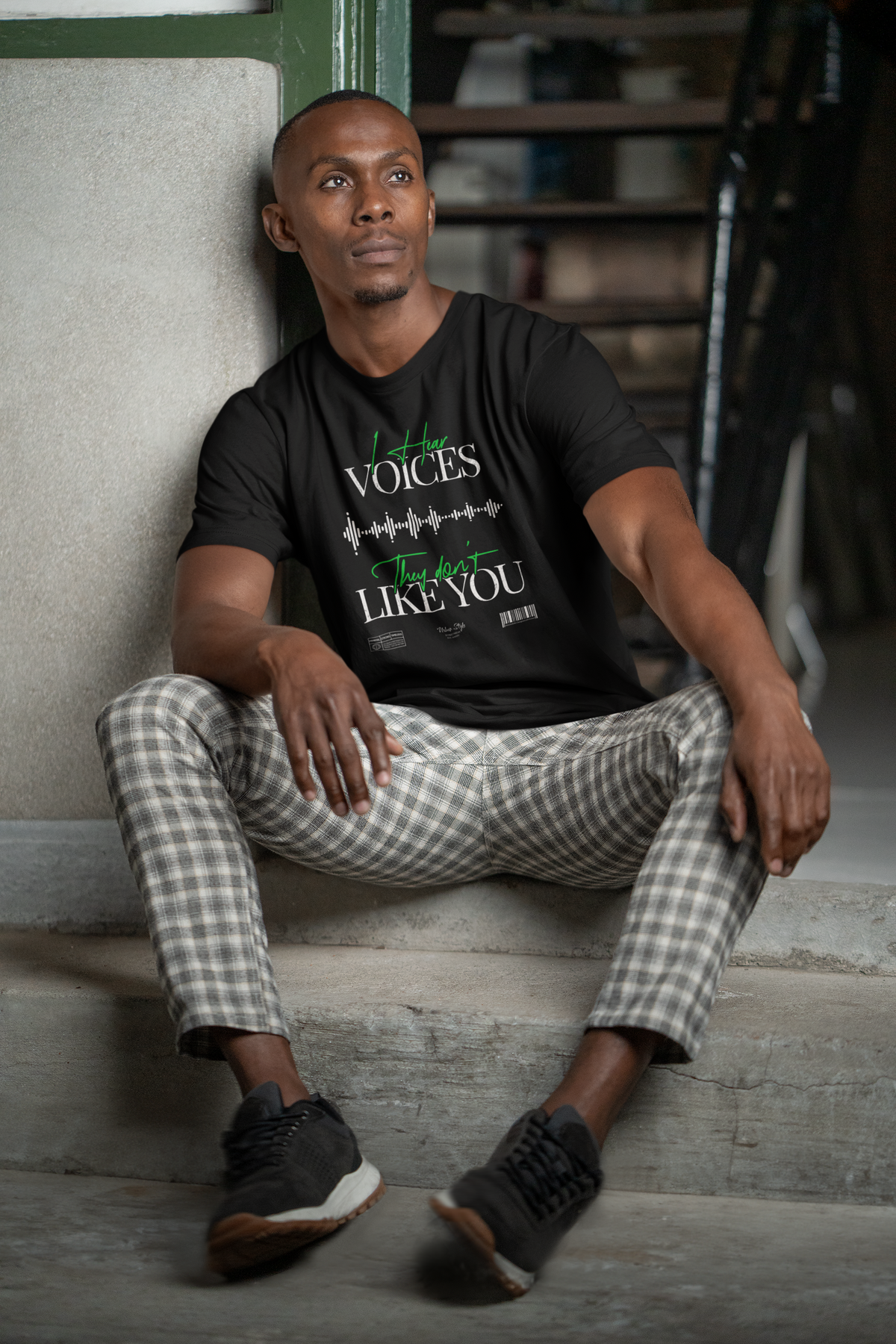 A seated male model wearing the "I Hear Voices / They Don’t Like You" black t-shirt by KRASSWERK, leaning casually against a stairwell wall in checkered pants — design shows glitchy waveform and green handwritten text.
