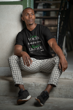 A seated male model wearing the "I Hear Voices / They Don’t Like You" black t-shirt by KRASSWERK, leaning casually against a stairwell wall in checkered pants — design shows glitchy waveform and green handwritten text.