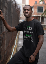 A serious male model wearing the "I Hear Voices / They Don’t Like You" black t-shirt by KRASSWERK, leaning against a wire fence in an urban alley — the design shows a distorted waveform and bold white-green typography.