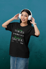 A smiling female model wearing the "I Hear Voices / They Don’t Like You" black t-shirt by KRASSWERK, standing against a teal wall with headphones on — the design features a glitch waveform and vivid green handwriting.