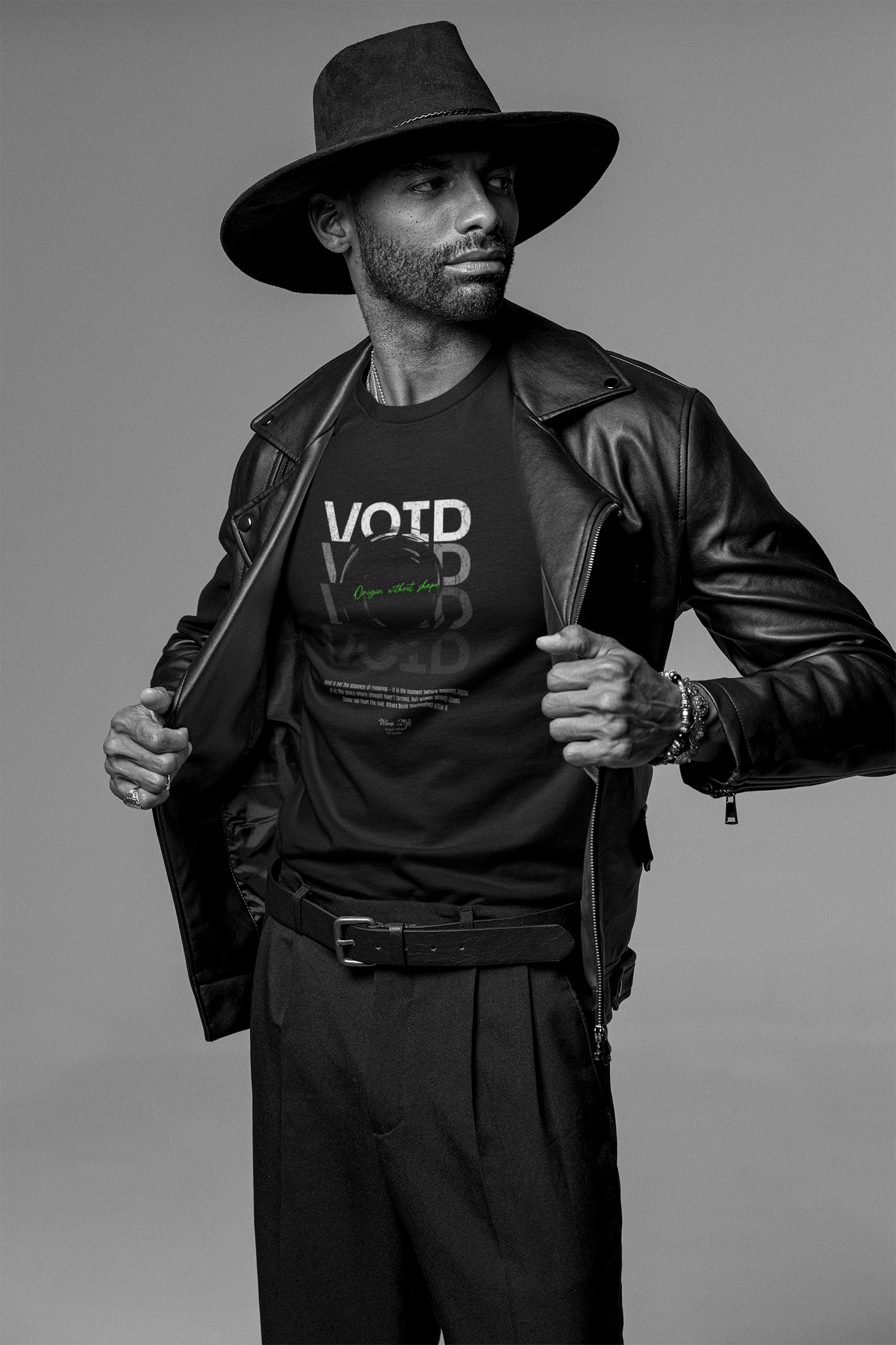 A confident male model in a black leather jacket and wide-brim hat pulls open his coat to reveal the VOID t-shirt. The design features layered typography and a green-glowing black hole at the center, symbolizing quiet collapse. Shot in a minimal studio with soft lighting.