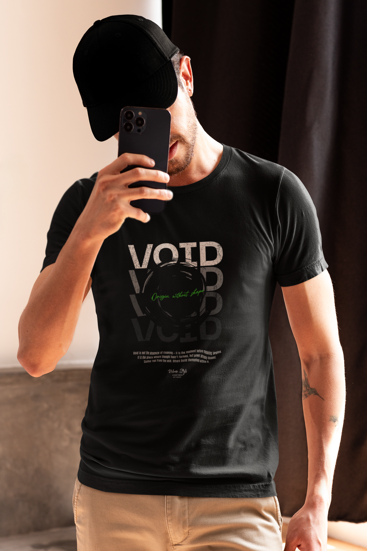 A man stands in soft light, partially hidden behind his phone. The VOID t-shirt features layered reflective text and a black hole at the center. The design whispers solitude and depth — a silent tribute to those who stare into the dark and don’t flinch.