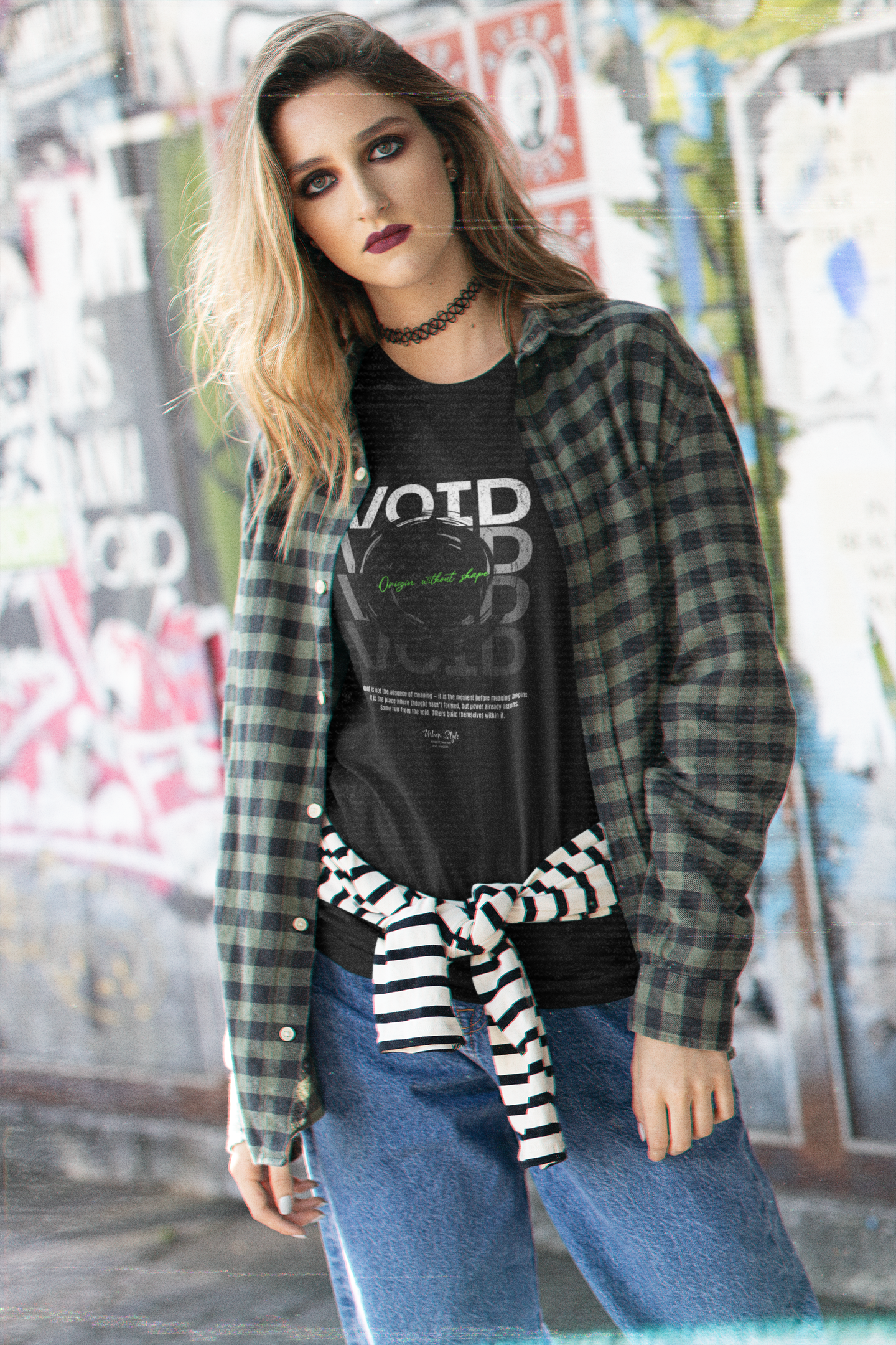 A young woman stands in front of graffiti-covered walls, wearing a plaid shirt over the VOID t-shirt. Her gaze is bold, her style grunge. The shirt features a glowing green message over a black hole design, symbolizing silent collapse and introspection.
