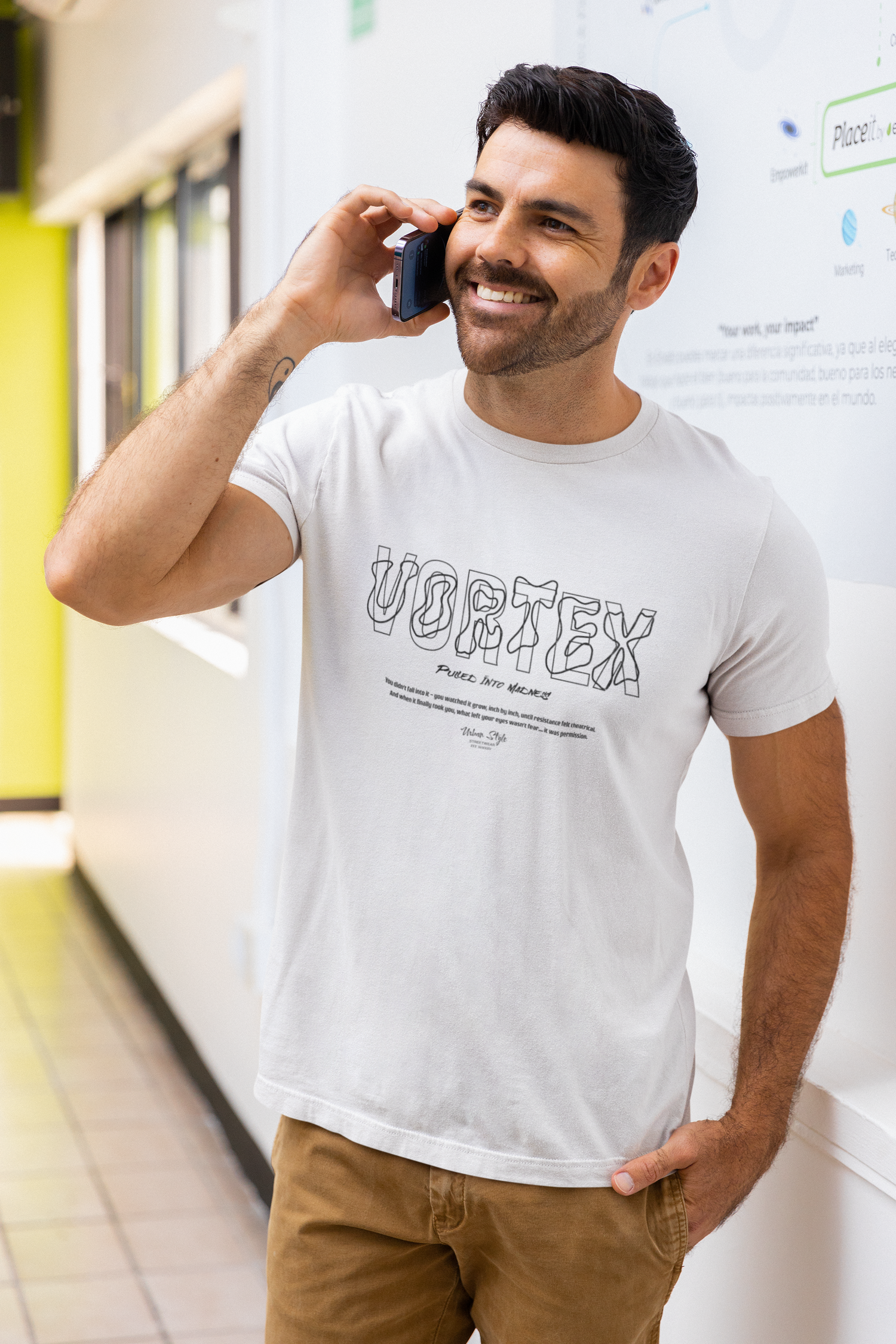 A smiling man taking a phone call in a bright hallway, wearing the white “VORTEX – Pulled Into Madness” t-shirt. The design features hand-drawn 3D lettering and a reflective quote about mental spiral and silent surrender.