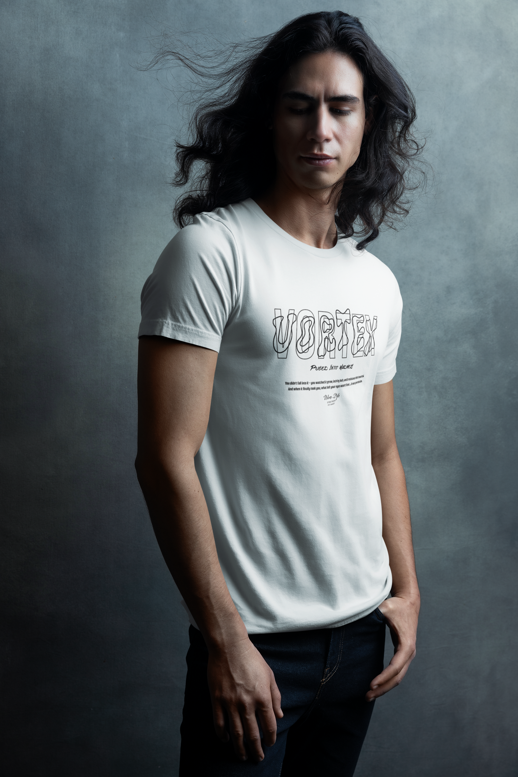 A man with long flowing hair stands against a moody textured backdrop, eyes closed and hands relaxed, wearing the “VORTEX – Pulled Into Madness” t-shirt. The surreal 3D line art typography mirrors the quiet intensity of the moment.