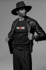 A man in a wide-brim hat and leather jacket poses confidently, revealing the black “WILDTHING” t-shirt with bold red claw marks and the phrase “100% INSTINCT.” Monochrome studio lighting adds intensity to the scene.