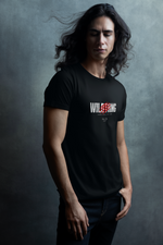 A man with long flowing hair stands calmly in front of a moody textured backdrop, wearing the black “WILDTHING” t-shirt featuring red claw marks and distressed typography. The bold design contrasts with the soft light and serene pose.