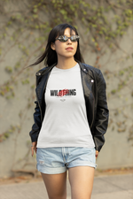 A woman walks confidently outdoors in a white “WILDTHING” t-shirt, featuring bold typography and red claw marks. She wears a black leather jacket and sunglasses, with vines and a concrete wall in the background.