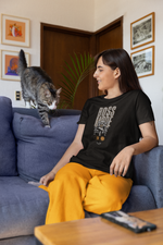 A calm female model lounges on a couch in cozy homewear, wearing the BOSS MODE tee as a curious cat steps over. The shirt shows a confident figure on a throne with the phrase “BOSS MODE” and a glowing ON switch.